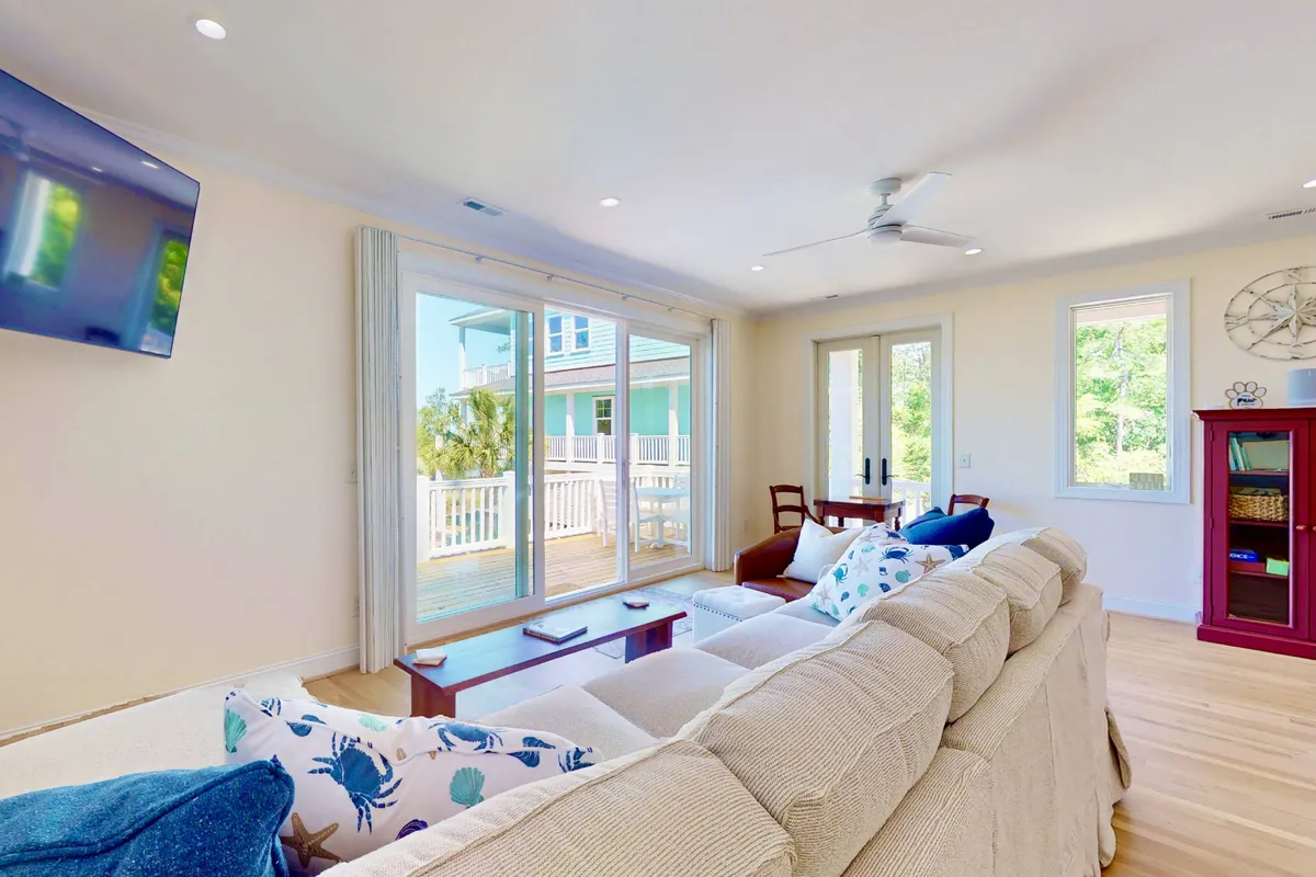 A comfortable living room featuring a plush sofa (ideal for use as a sleeper), large sliding glass doors bringing in natural light, a colorful array of pillows, and a decorative wall clock. The space is casual, welcoming, and perfect for family or friends needing flexible sleeping arrangements.