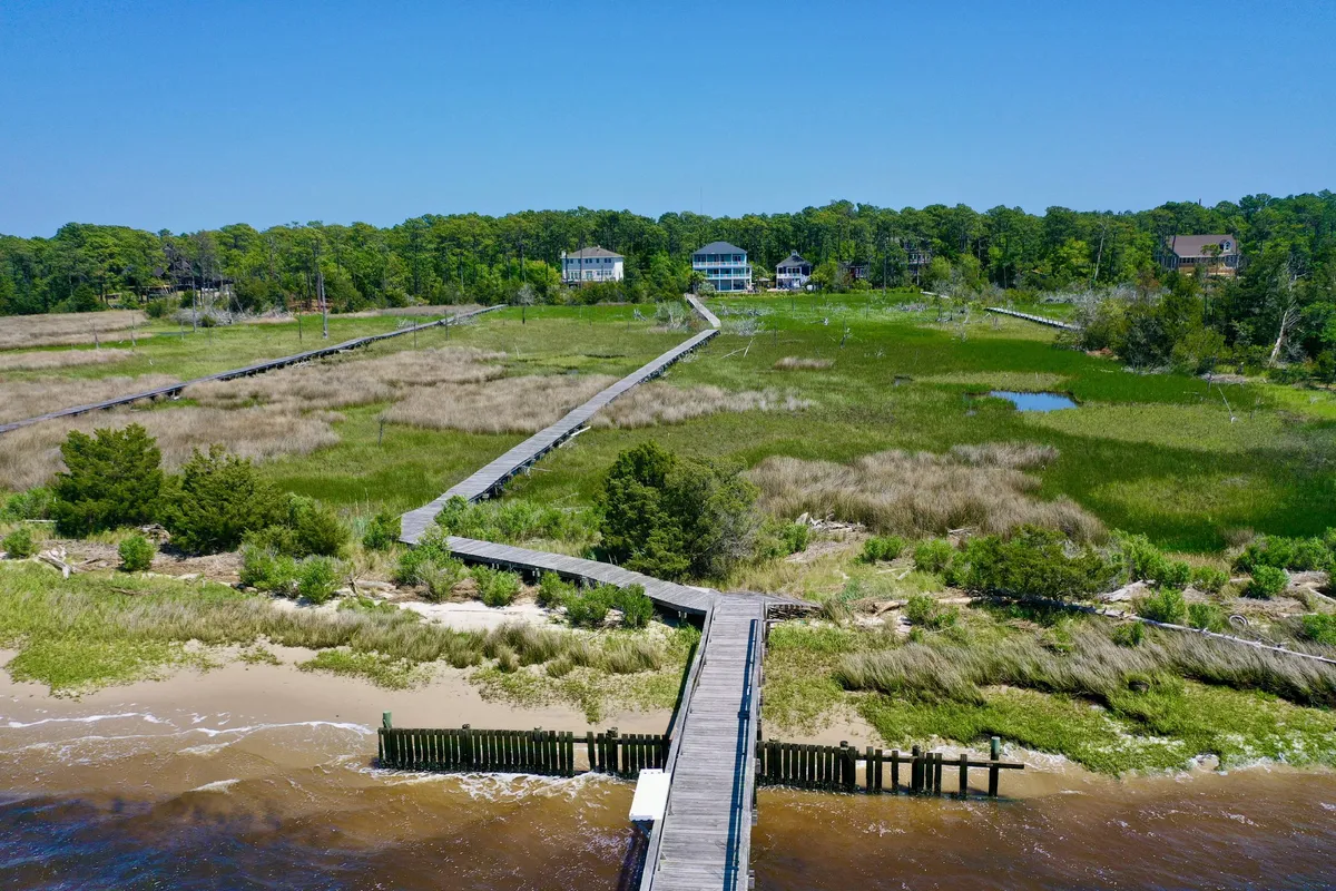 A picturesque wooden boardwalk meanders through lush green marshes, with waterfront houses in the distance. The area is quiet, ideal for walking, bird watching, and taking in the peaceful environment. The boardwalk is welcoming for both people and pets, offering a safe, scenic route for exploration.