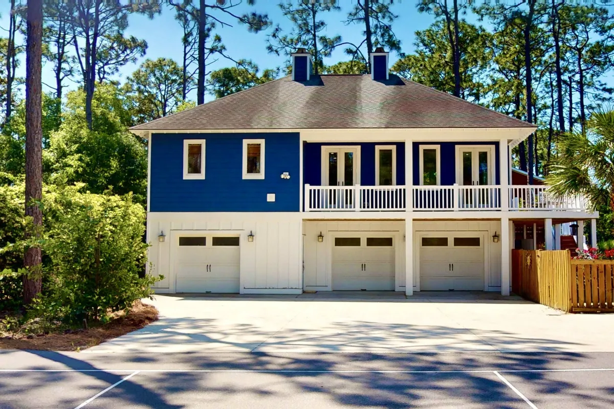A bright, cheerful exterior view of the blue and white home, surrounded by mature trees and featuring a balcony and garage doors. The ample outdoor space offers room for pets to roam and for families to gather, with the serene surroundings tying the home to the natural landscape.