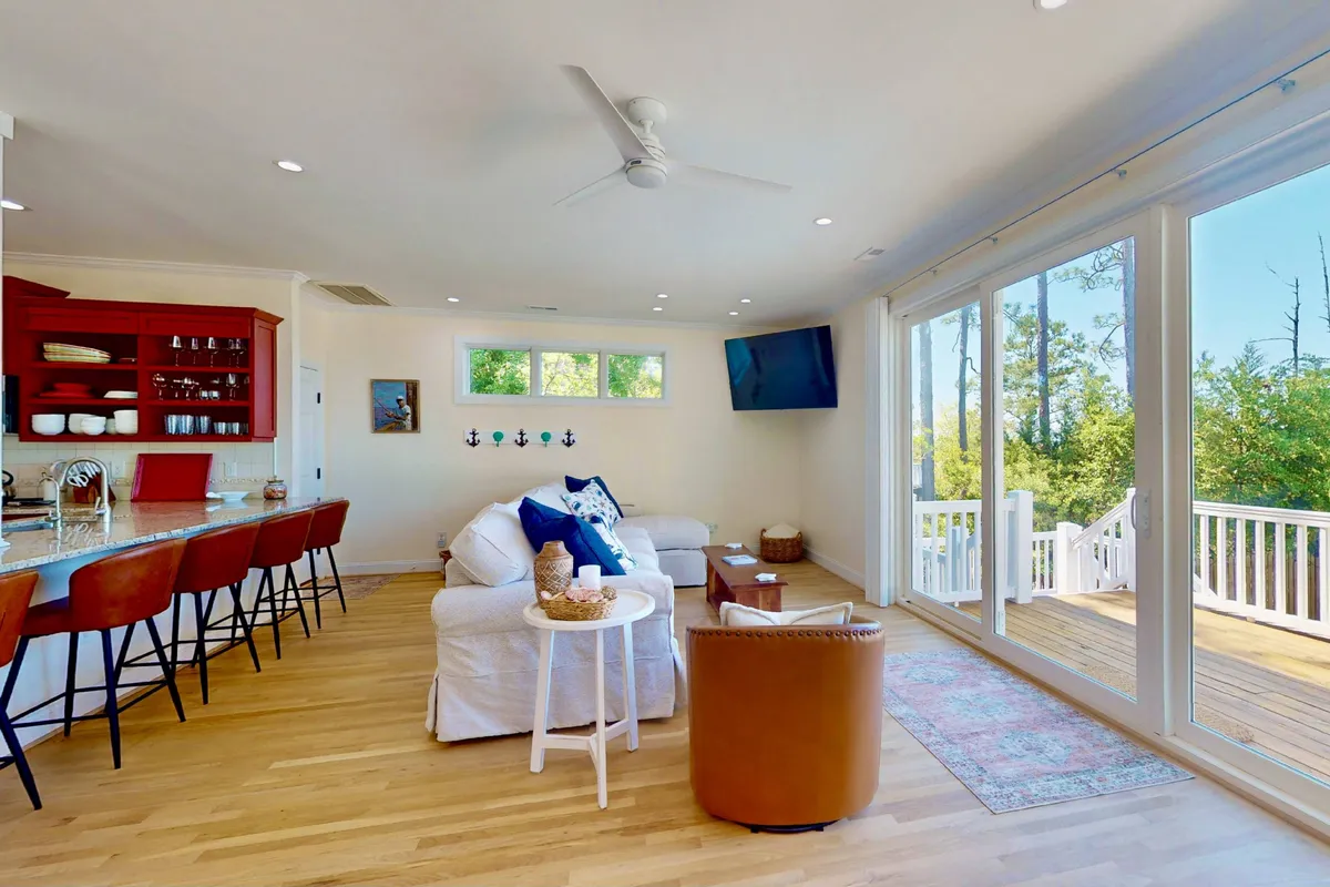 A cozy living space with a large sofa and kitchen bar area, complemented by decorative wall art and expansive windows offering lovely outdoor views. The area is filled with natural light and designed for gatherings and relaxation.