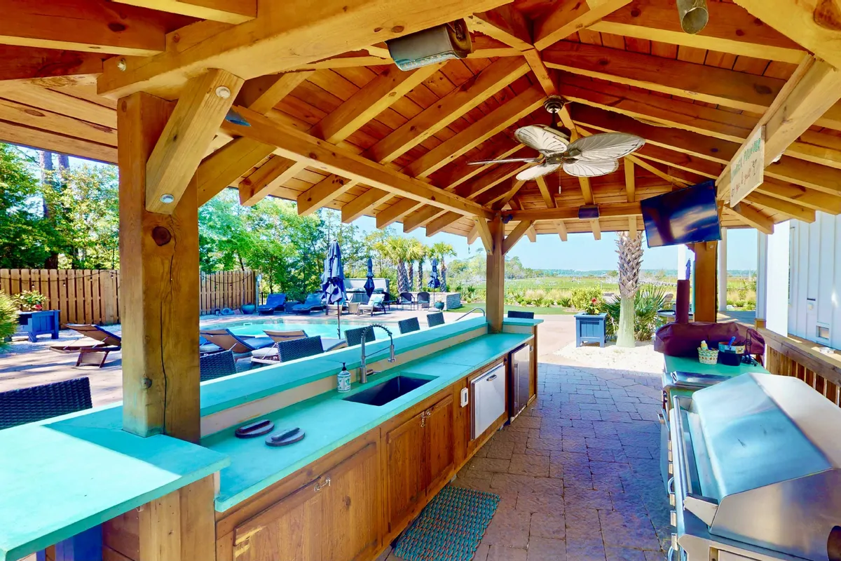 A vibrant outdoor kitchen and barbecue area adjacent to a sparkling swimming pool. The scene includes a contemporary grill, a bar-style seating area, overhead fans, and comfortable lounge chairs, ideal for entertaining or family gatherings.