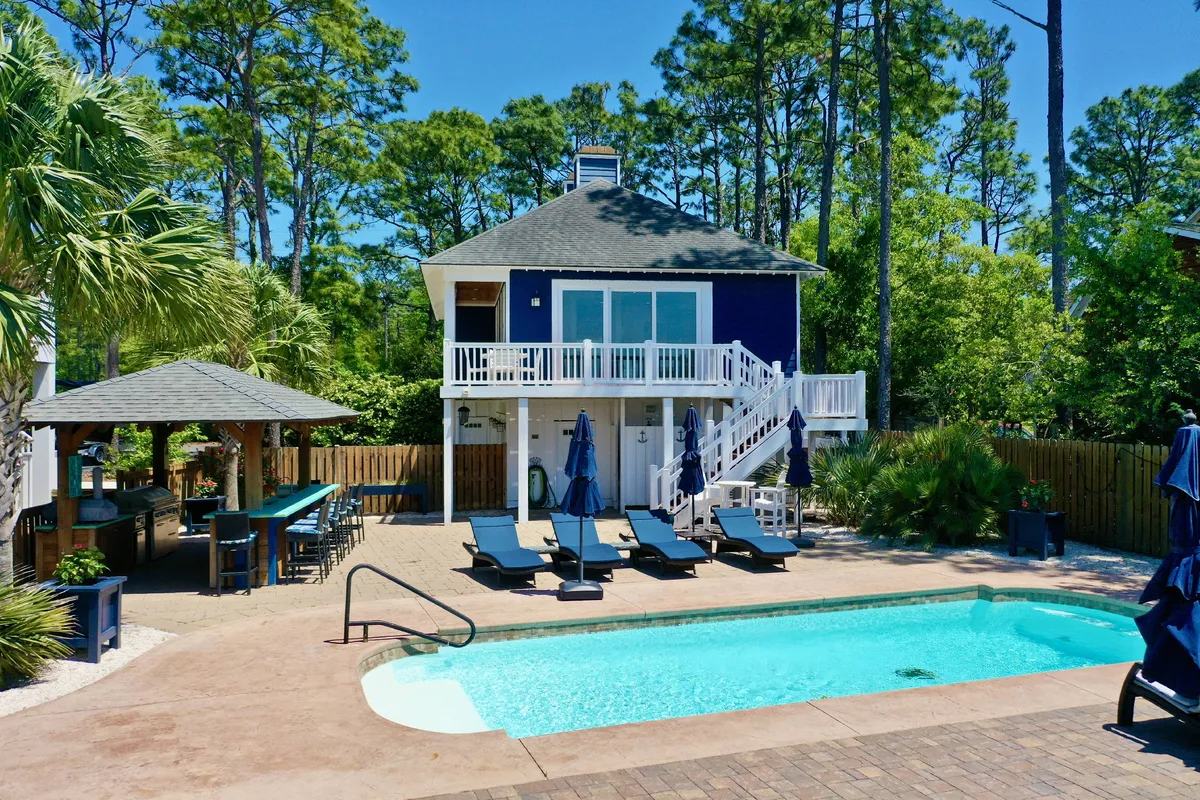 The image showcases a vibrant blue single-family house in Wilmington, NC, set amidst lush greenery. The inviting swimming pool is surrounded by comfortable outdoor seating, perfect for relaxation or social gatherings. The scene suggests opportunities for sunbathing, swimming, and evening barbecues, all under a relaxed, welcoming atmosphere.