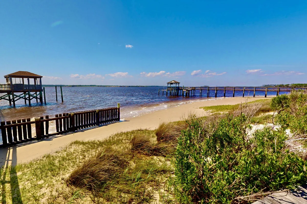 Wooden piers stretch out from a sandy beach framed by grassy dunes, with gentle waves lapping at the shore. The view is expansive, evoking the coastal setting and nearby recreational opportunities for families and kids.