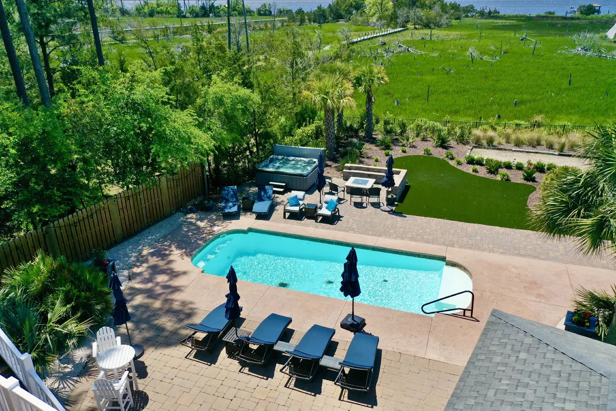 A sun-drenched swimming pool area featuring a relaxing hot tub, inviting lounge chairs, and a lush green lawn bordered by mature trees. The setting exudes tranquility, offering both sun and shade for poolside relaxation. The pool and hot tub are central, suggesting a seamless transition from swimming to soaking, perfect for all ages.