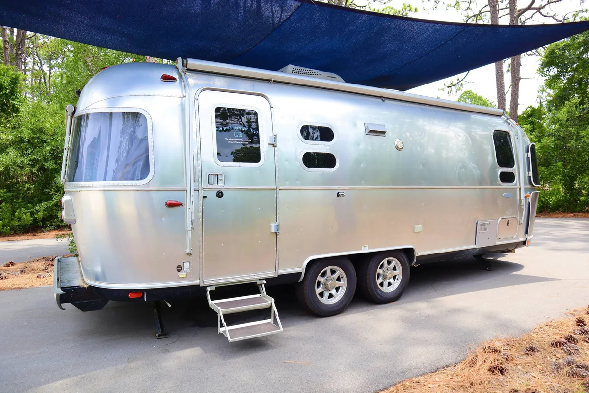 This exterior image features the Airstream trailer set in a shaded area surrounded by greenery, clearly highlighting its standalone placement and connection to nature. The Airstream is shown as a separate, private guest suite just steps from the main home, embodying both the adventure and independence aspects described.