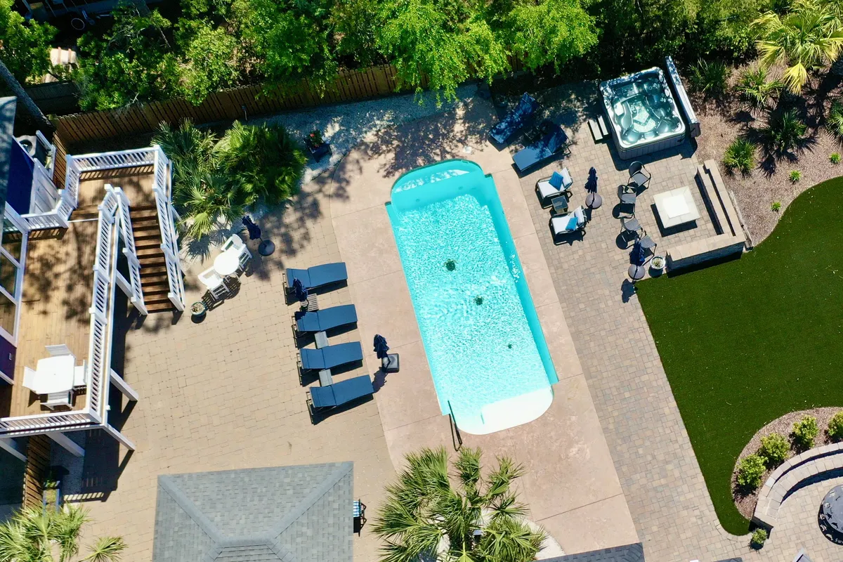A sunlit view of the pool and hot tub with inviting lounge chairs and a spacious deck area, surrounded by lush greenery. The scene exudes relaxation and sets the stage for social gatherings by the water, with ample space for groups to unwind together.