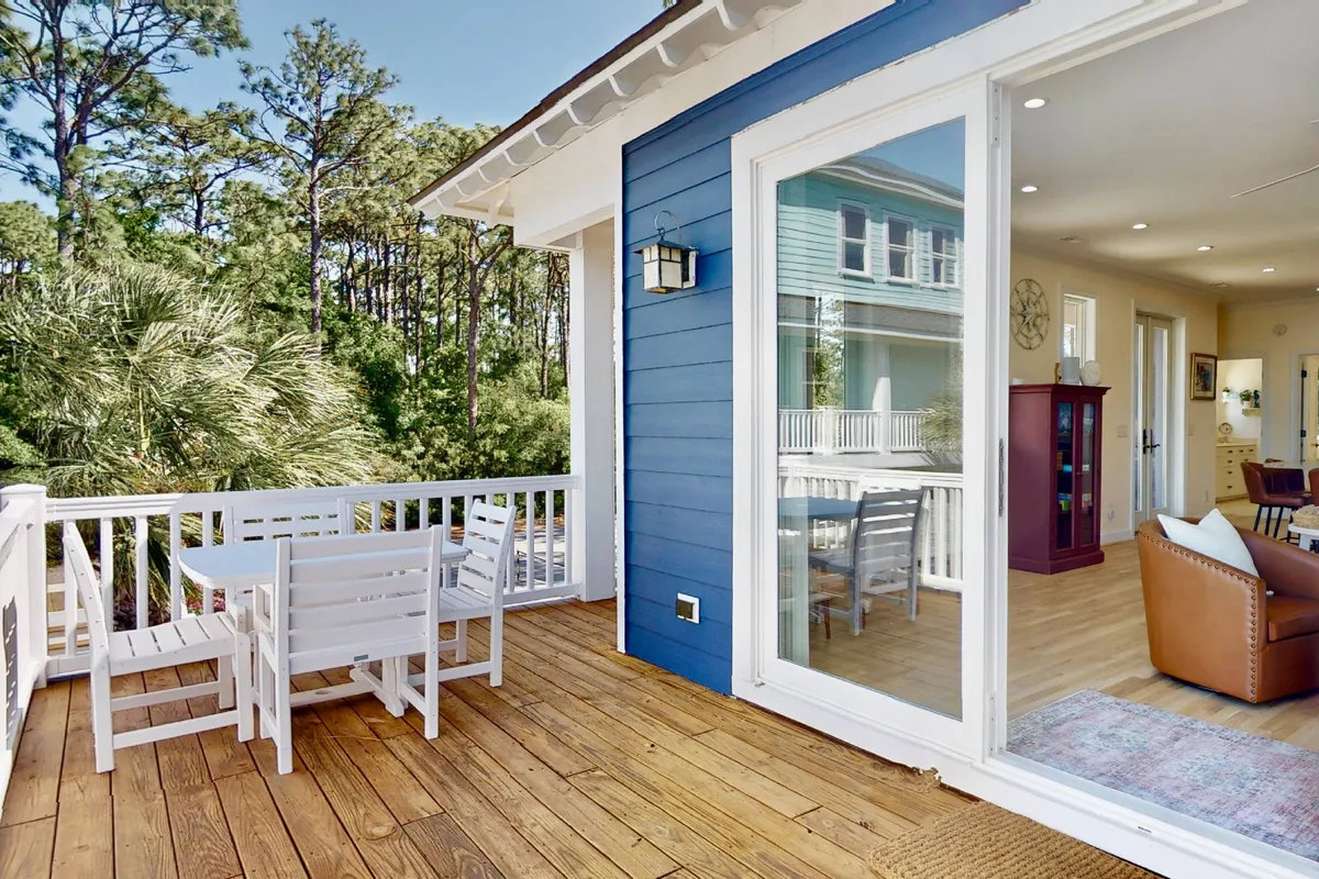 This image features a beautiful outdoor wooden deck furnished with white chairs and surrounded by green trees, adjacent to a striking blue house exterior. The tranquil setting highlights spaces for outdoor dining, relaxing, and enjoying the property's natural environment.