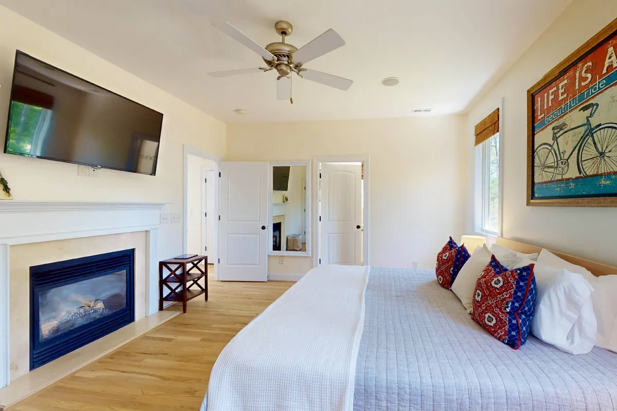 A tranquil bedroom anchored by a king-sized bed dressed in fresh, plush linens. Decorative pillows, a warm fireplace, and a large TV create a cozy, inviting environment, while playful wall art adds charm. Sunlight filters through the window, highlighting a space ideal for relaxation or winding down after adventure-filled days.
